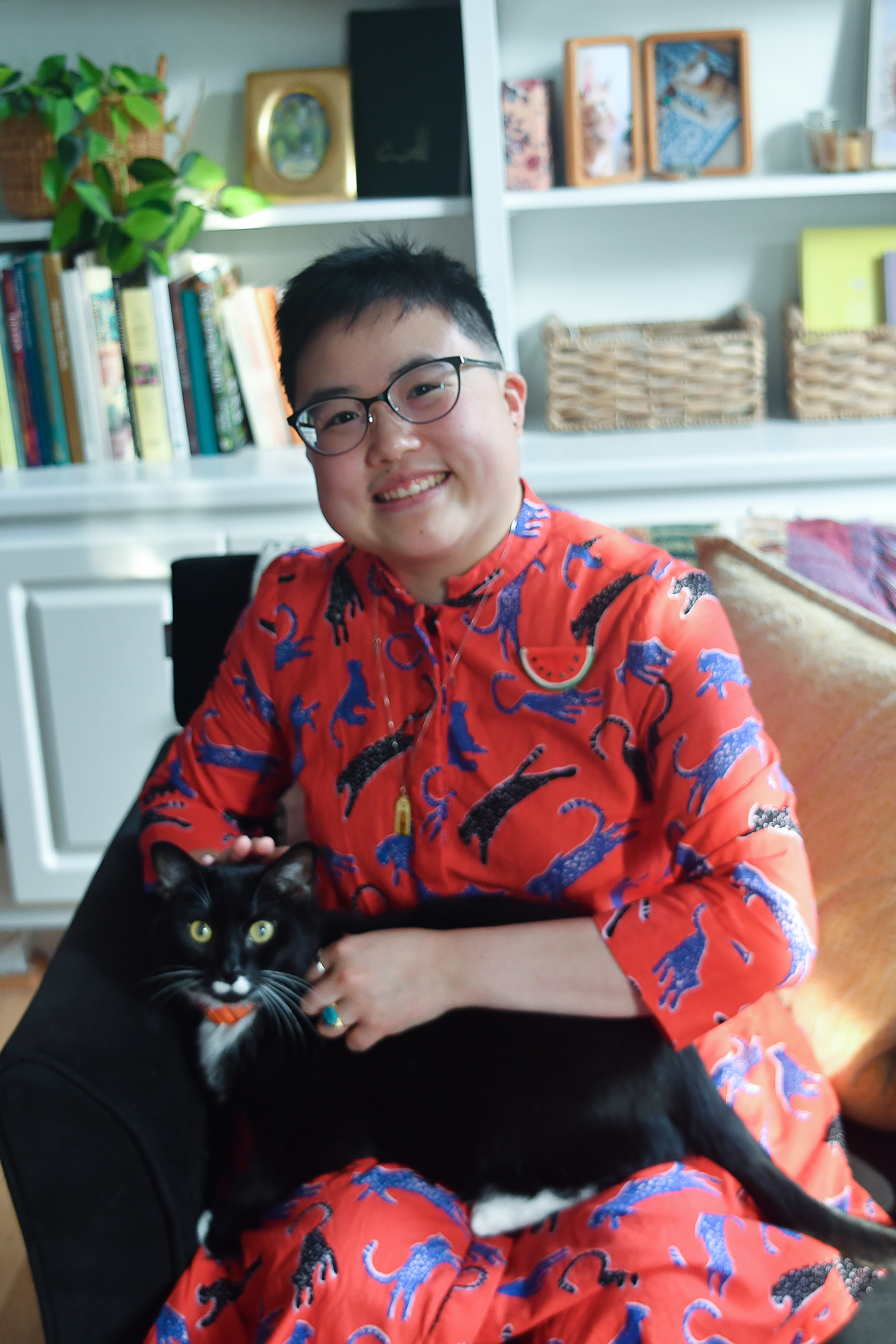 Ly Xīnzhèn smiles while sitting in a tan leather chair, wearing a red outfit and holding a black tuxedo cat. There are bookshelves in the background.
